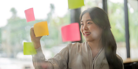 Smiling businesswoman Asian people thinking and use sticky notes on glass wall in officeの写真素材