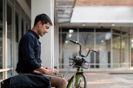 Asian businessman with bicycle using laptop and sitting outside the office building. man commuting...