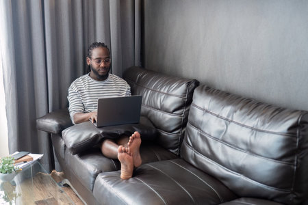 Young African American man holding laptop relaxing sitting on sofa working remote at homeの写真素材