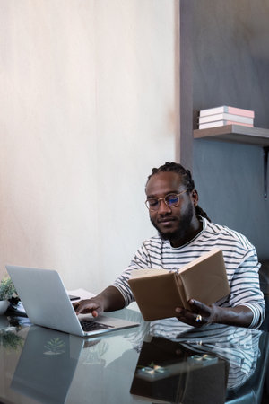 Young african man using laptop at home, black male looking at read book relaxing on leisure with...