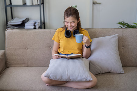 Young Woman Reading a Book at Home on a Comfortable Sofa with a Cup of Coffee, Relaxing and Enjoying...