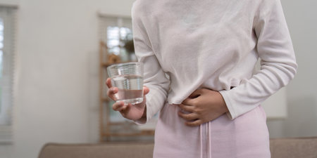 Woman Holding Stomach in Pain While Holding a Glass of Water, Depicting Abdominal Discomfort and...