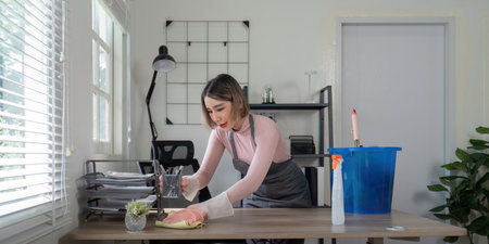 Young Maid Cleaning Modern Office Desk with Cleaning Supplies in Bright Room