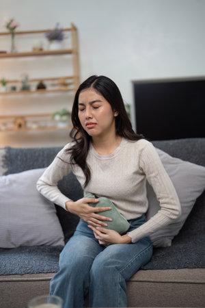 Young Woman Experiencing Abdominal Pain While Sitting on a Couch at Home, Holding a Hot Water Bottle for Reliefの写真素材