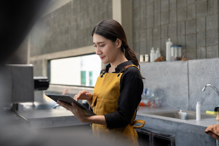 Young Barista in Yellow Apron Using Tablet in Modern Coffee Shopの写真素材