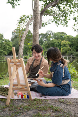 Romantic Picnic Couple Drawing Outdoors in Nature with Easel and Painting, Capturing Creative Moments in a Serene Park Settingの写真素材