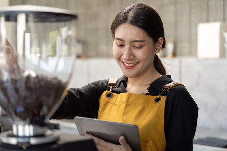 Smiling Barista in Yellow Apron Using Tablet in Modern Coffee Shopの写真素材