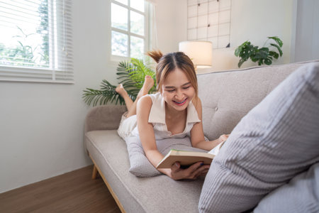Young Woman Relaxing on Sofa Reading a Book in Bright Modern Living Room with Natural Lightの写真素材