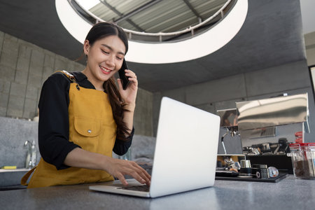 Young Woman Barista Working on Laptop and Talking on Phone in Modern Coffee Shopの写真素材