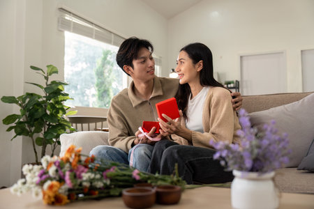 Happy Couple Exchanging Gifts on Sofa in Cozy Living Room with Flowers and Natural Lightの写真素材