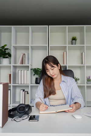 Young Student Studying Online at Home Desk with Books and Headphones, Taking Notes in Notebookの写真素材