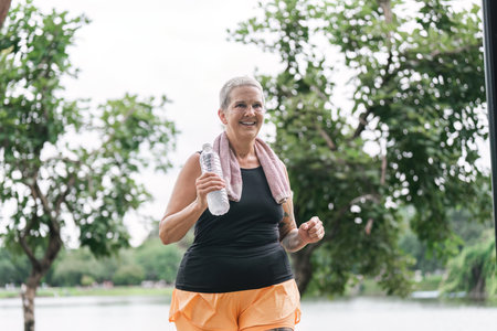 Joyful Caucasian senior woman jogging in summer park with waterの写真素材