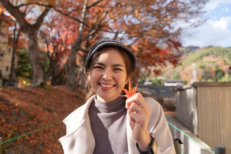 Smiling Asian Woman Enjoying Autumn Holding a Maple Leafの写真素材