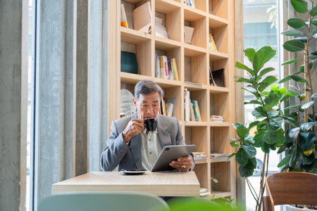 Elderly man sipping coffee while reading a tablet in a vibrant cafe filled with greeneryの写真素材
