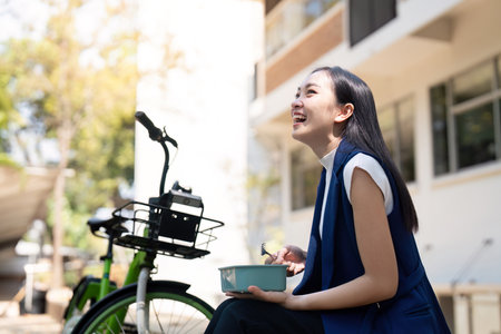Joyful businesswoman enjoying a meal outdoors beside her eco-friendly bikeの写真素材