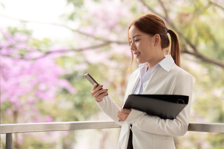 Engaged businesswoman using smartphone while holding files against a backdrop of blooming flowersの写真素材