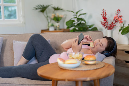 Overweight woman relaxing on the couch, enjoying snacks while scrolling on her phone at homeの写真素材
