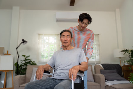 Son assisting elderly father in a wheelchair within a bright living room.の写真素材