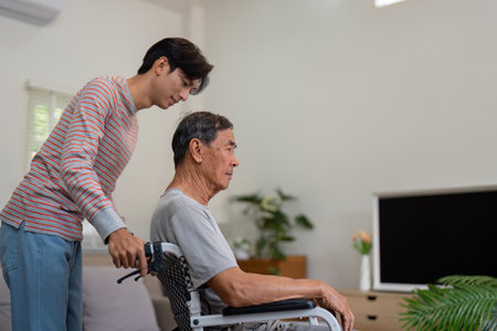 Son pushing elderly father in a wheelchair through a bright living room.の写真素材