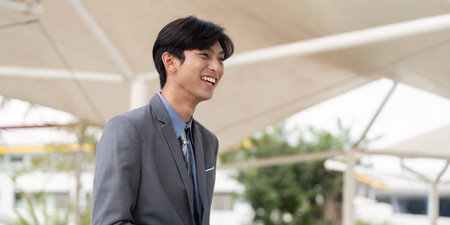 Smiling young Asian businessman in a suit enjoying a moment outdoors in a modern business environment.の写真素材