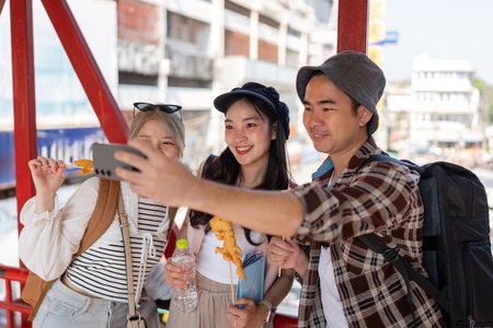 Young friends taking a selfie while enjoying street food at a vibrant market, celebrating travel and friendship.の写真素材