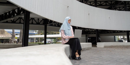 Focused Muslim businesswoman working on a laptop in a modern outdoor setting. Emphasizing productivity and cultural diversity in entrepreneurship.の写真素材