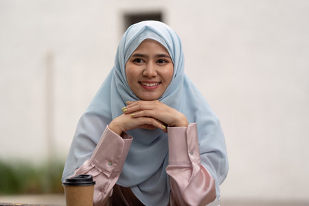 Businesswoman in hijab smiling at camera with coffee cup in outdoor setting.