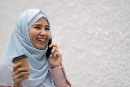 Smiling Muslim businesswoman talking on the phone while holding a coffee cup. Concept of modern...
