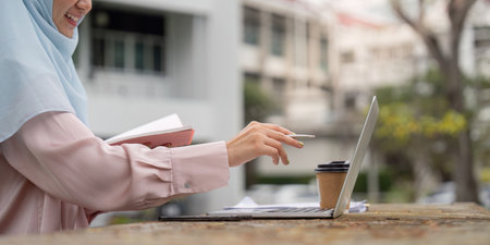 Focused Muslim woman using a laptop and taking notes outdoors, emphasizing productivity and organization.の写真素材