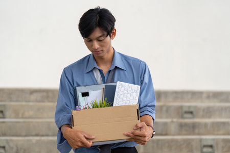 Young man leaving office with personal belongings in a box after resignation, reflecting on career transition.の写真素材