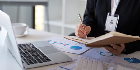 Financial professional taking notes and analyzing data on a laptop at a modern office desk.の写真素材