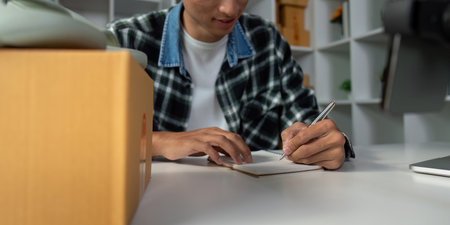 Young entrepreneur taking notes for online sales strategy, writing in notebook at modern workspace...