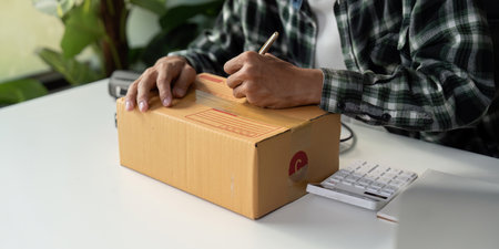 Person writing on cardboard box while preparing for online order shipment with calculator nearby.