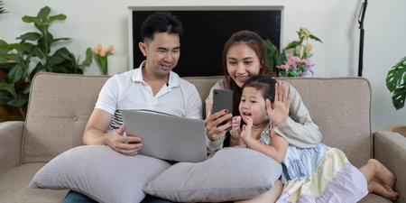 Family video calling together on a couch, sharing smiles and waves with a smartphone.の写真素材