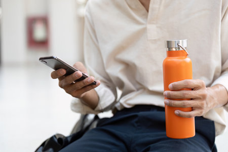 Eco-Conscious Commuting and Hydration. A fashionable young man with a reusable water bottle reaches for his smartphone, embodying a modern, sustainable lifestyle.の写真素材