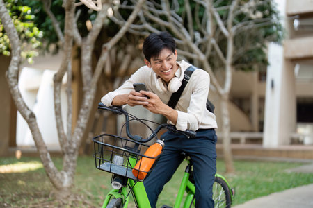 Communication and Joy. A young man sitting on a bike, smiling while checking his smartphone outdoors in a lively setting.の写真素材