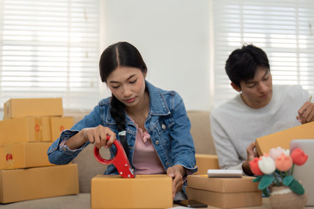 Preparation and Execution. A young woman meticulously packages items while her partner prepares for their next business task.の写真素材