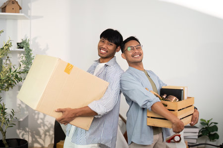Joyful gay couple carrying boxes, celebrating their new beginning togetherの写真素材