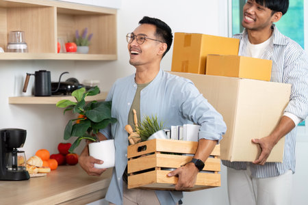 Gay couple moving in together, carrying boxes and plants in a cozy kitchenの写真素材