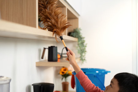 Child Helping with Cleaning. Young boy dusting shelves with a feather duster in a bright kitchen.