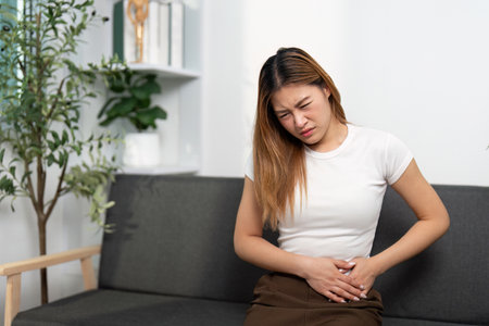 Health Awareness: Young woman experiencing abdominal pain while sitting on a couch.の写真素材