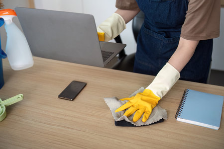 Home Cleaning. Young woman with gloves cleaning her desk while using a laptop.