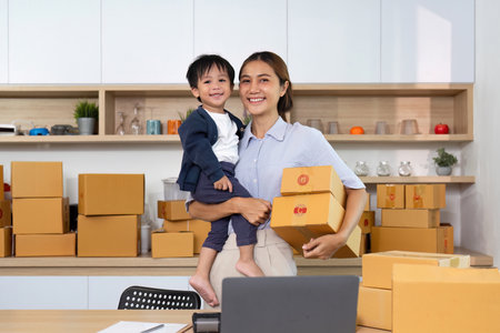 Happy mother holding boxes with son in home officeの写真素材