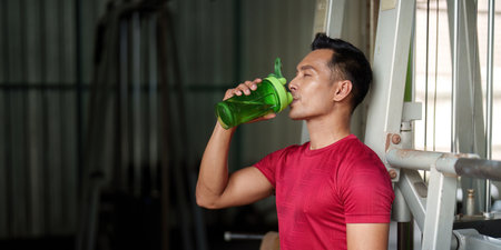 Hydration. Young man drinking from shaker bottle after workout in gym.