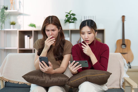 Surprised Reactions. Two friends looking shocked while checking their phones in a cozy living room.の写真素材