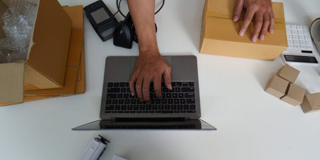 E-commerce Workflow. Overhead view of young man typing on laptop while handling packages.