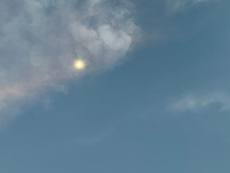 A calm blue sky with the sun softly glowing behind thin clouds, creating a quiet and dreamy...