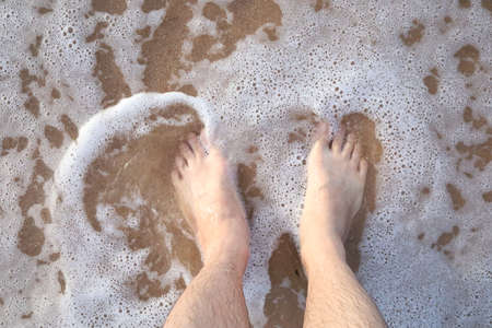 Top view of male feet on sandy beach with foamy waves.の写真素材