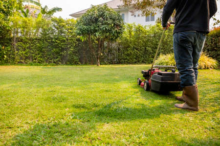 A worker man cutting grass in green yard with electric lawnmower on sunlight.  Man with electric...