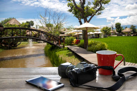red cup of coffee with retro camera and mobile phone on wooden table in nature background, holiday conceptの写真素材